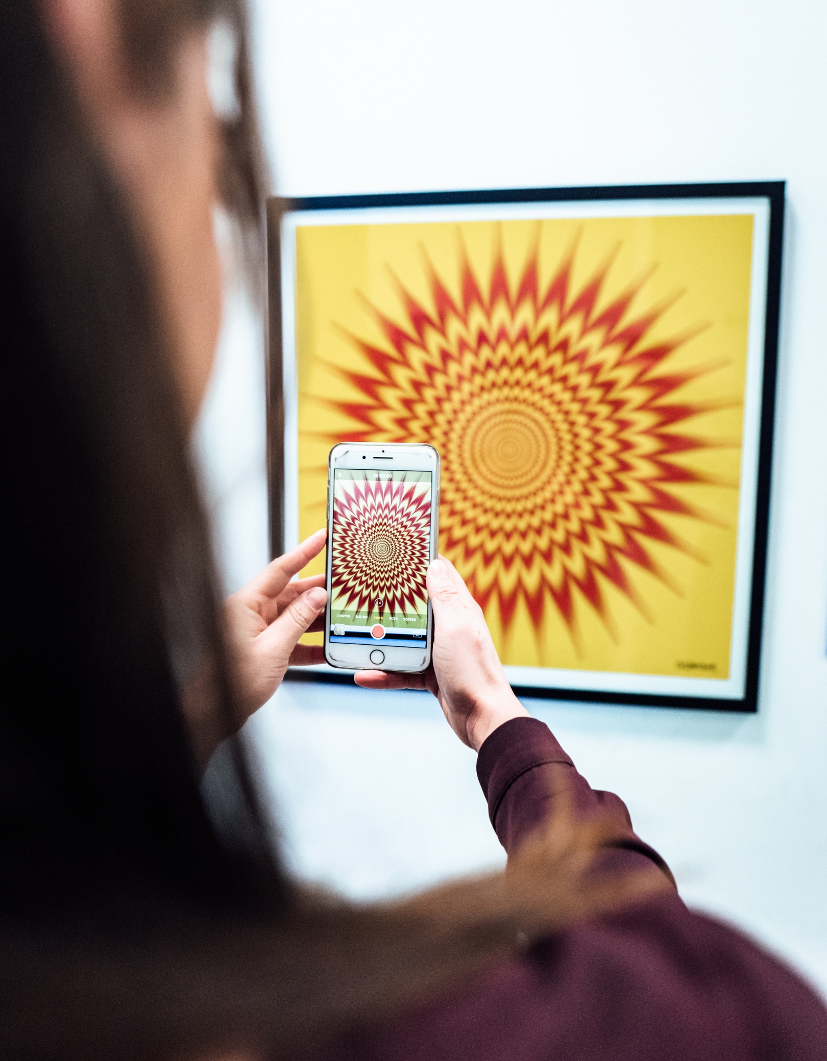 Eine Frau hält ein Smartphone und fotografiert ein gerahmtes Kunstwerk mit einer gelb-roten optischen Illusion im Museum der Illusionen Hamburg; das Bild zeigt einen spiralförmigen, pulsierenden Effekt.