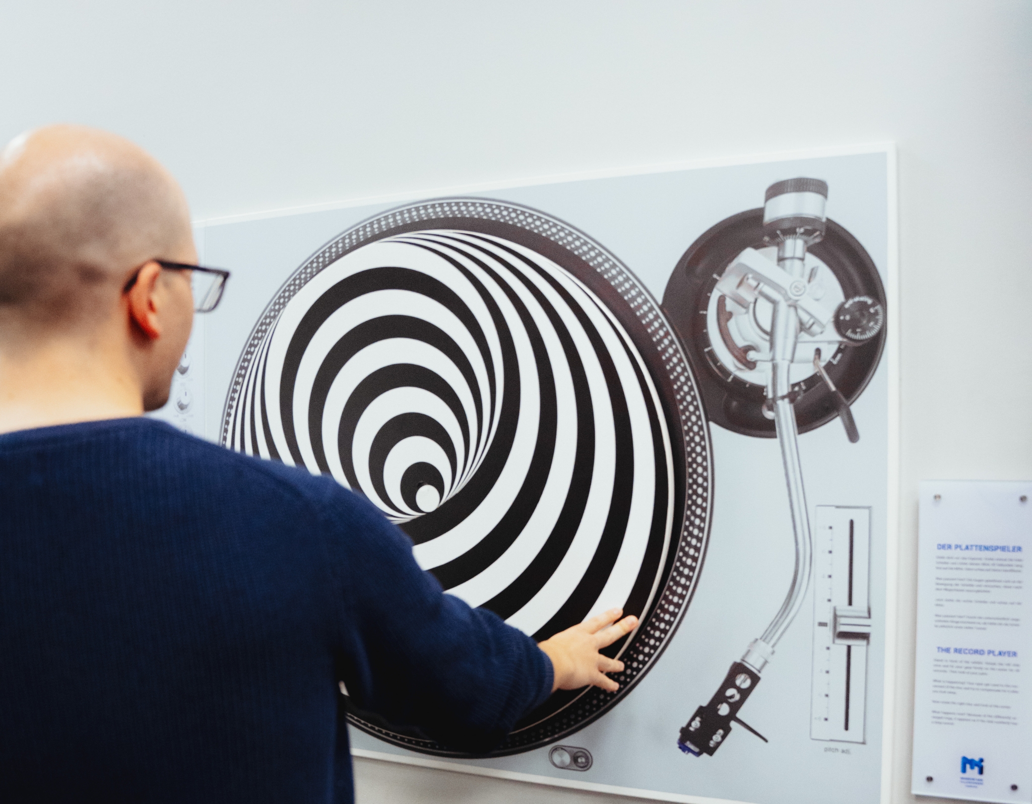 Ein Mann betrachtet und berührt eine interaktive Illusion in Form eines Plattenspielers mit einem hypnotischen, schwarz-weißen Spiralmuster an der Wand im Museum der Illusionen Hamburg.