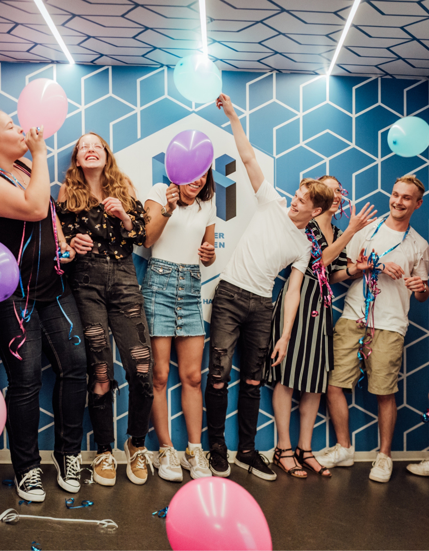 A cheerful group of young people celebrates with balloons and streamers in the Ames Room at the Museum of Illusions Hamburg.