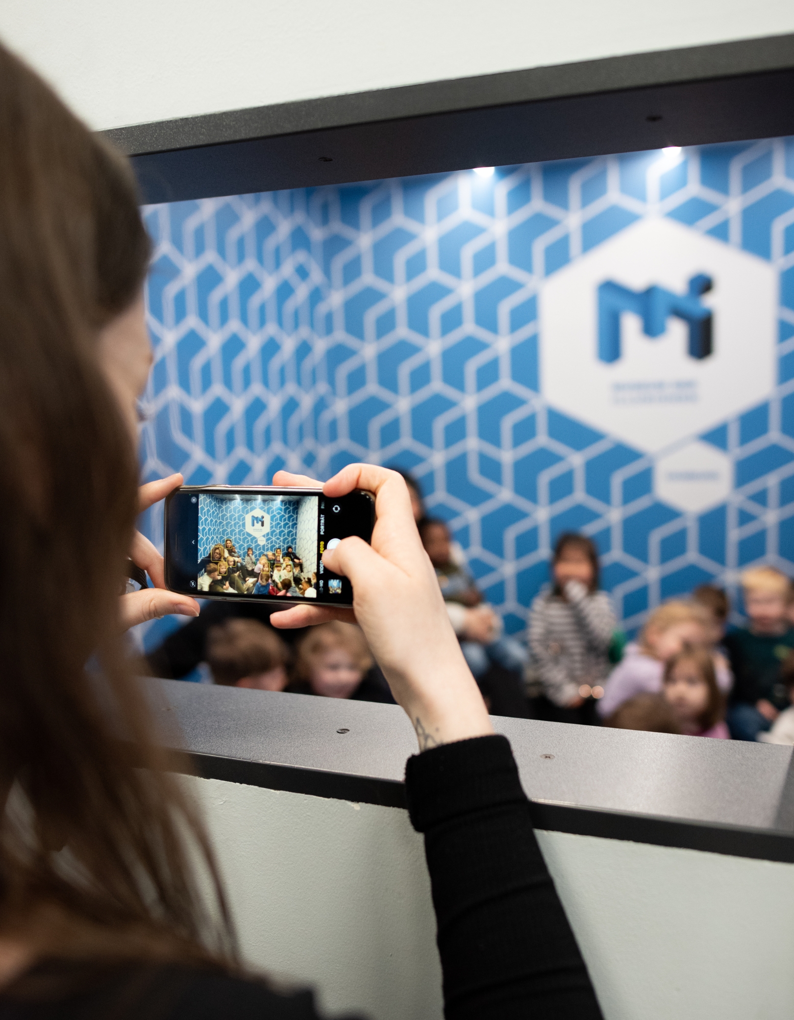 A woman takes a smartphone photo of a cheerful group of children in the Ames Room at the Museum of Illusions Hamburg.