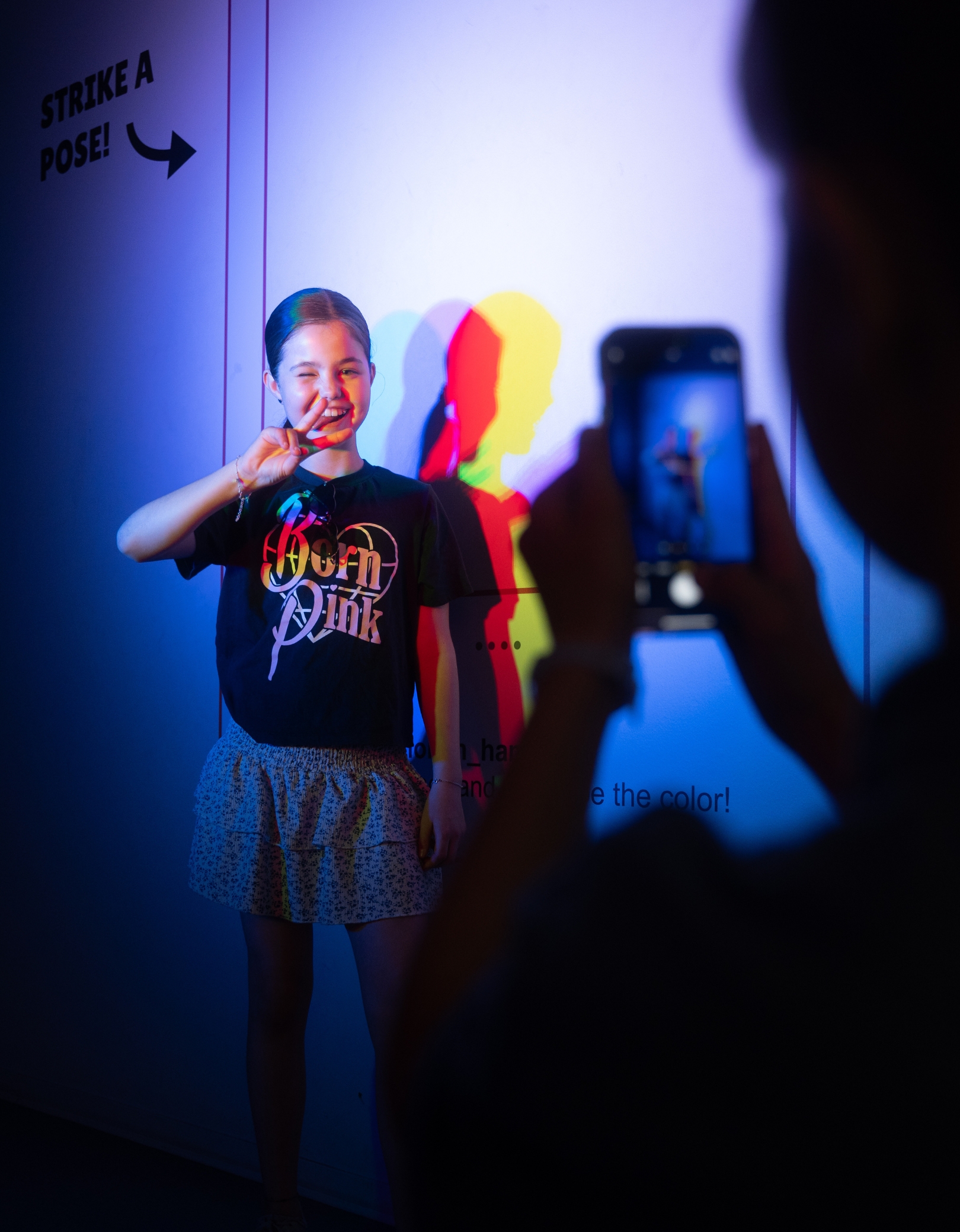 A girl poses with a peace sign in the Color Room at the Museum of Illusions Hamburg. Colorful lights cast multicolored shadows on the wall as someone takes a photo of her with a smartphone.