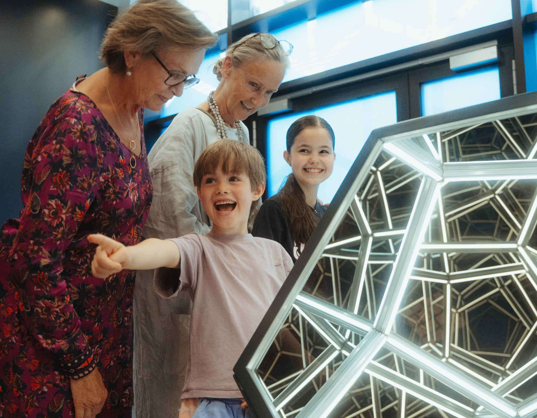 Zwei Kinder stehen mit ihren Großmüttern vor einer leuchtenden Licht- und Spiegelillusion im Museum der Illusionen 