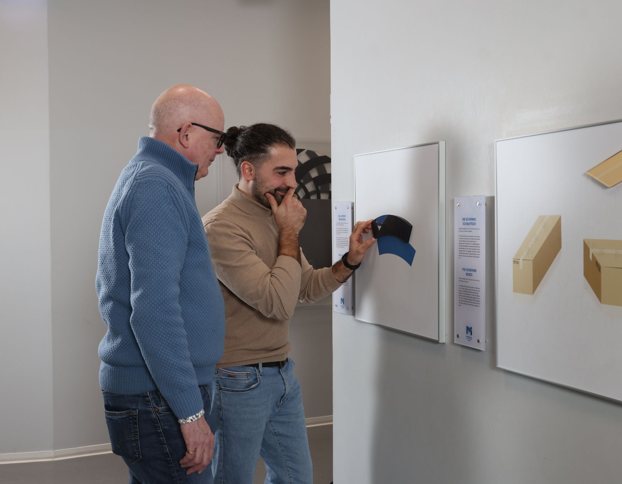 Zwei Männer stehen vor der Jastrow Illusionen und überlegen, wie die optische Illusion funktioniert.