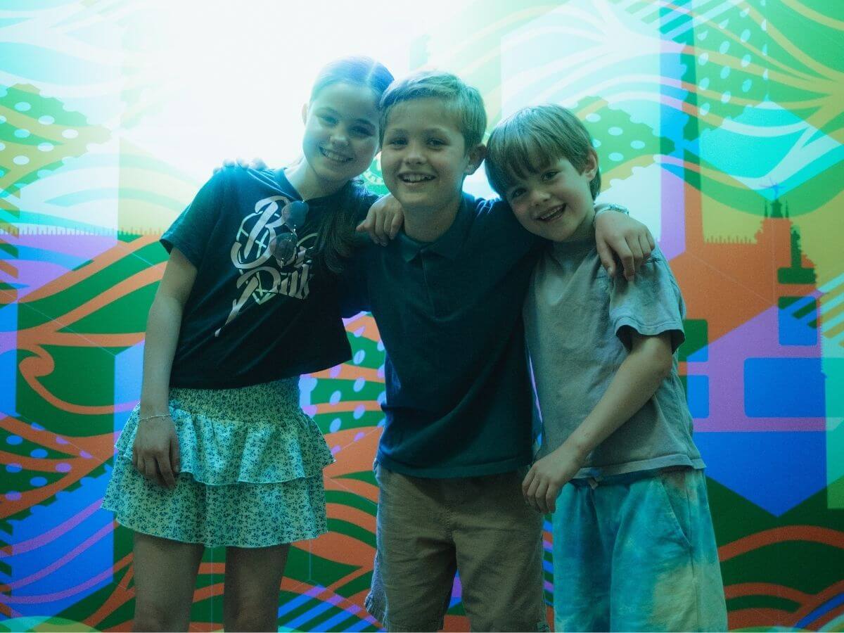 Drei Kinder stehen Arm in Arm in einem bunten Raum im Museum der Illusionen.