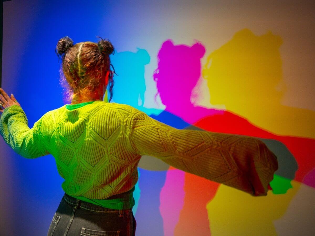 Ein Mädchen steht in einem Raum vor einer weißen Wand. Das Licht wird so gebrochen, das es bunte Schatten auf die Wand wirft.